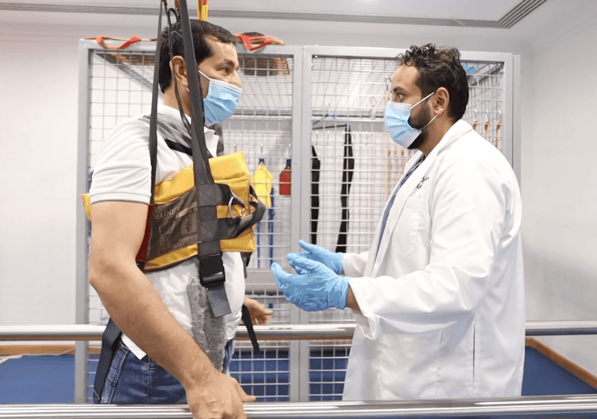 A man is suspended in a harness, engaging in conversation with Dr. Mina Gamil. Both wear face masks, and the doctor dons blue gloves. They are in a physiotherapy setting in Dubai, focusing on rehabilitation techniques for improved patient care.